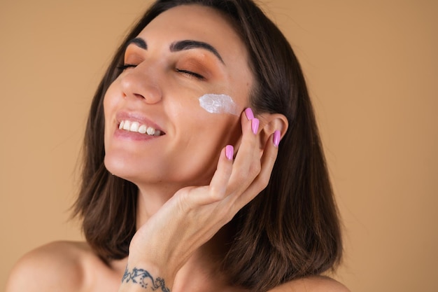 Бесплатное фото Портрет молодой женщины на бежевой стене с естественным теплым макияжем и гладкой чистой кожей, наносит крем на лицо