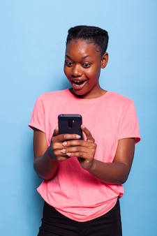 青い背景のスタジオに立っているスマートフォンを使用して友人とメッセージを送るアフリカ系アメリカ人のティーンエイジャーの肖像画。電話アプリを使用してソーシャルメディアを閲覧している若い女性。技術コンセプト