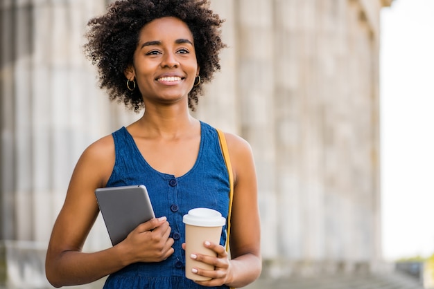 通りで屋外に立っている間デジタルタブレットとコーヒーを持っているアフロビジネス女性の肖像画 Premium写真