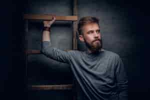 Бесплатное фото Портрет городского бородатого мужчины, позирующего возле старой винтажной лестницы на сером фоне.