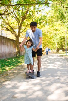 公園​で​スケート​ボード​を​している​アジア​の​父​と​彼​の​娘​の​肖像画​。​彼女​が​スケート​ボード​に​立っている​間​、​路地​を​歩いている​小さな​女の子​の​手​を​握って​幸せな​男​。​子供​と​父性​の​概念​と​の​アクティブな​休息