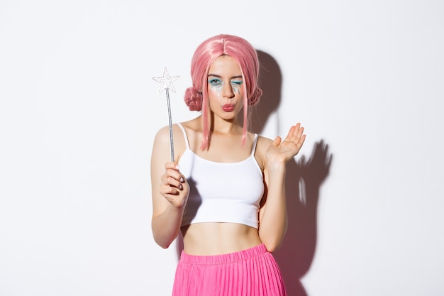 Портрет привлекательной девушки-феи косплей на хэллоуин, в розовом парике и держащей волшебную палочку, стоящую на белом фоне