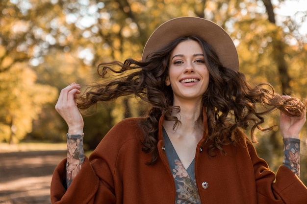 無料写真 暖かい茶色のコート秋の流行のファッション、帽子をかぶってストリートスタイルに身を包んだ公園を歩く長い巻き毛の魅力的なスタイリッシュな笑顔の女性の肖像画
