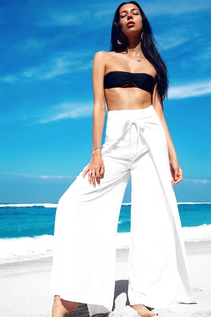 Бесплатное фото Портрет красивой кавказской модели женщины с темными длинными волосами в широких классических брюках, позирующих на летнем пляже с белым песком на голубом небе и океане