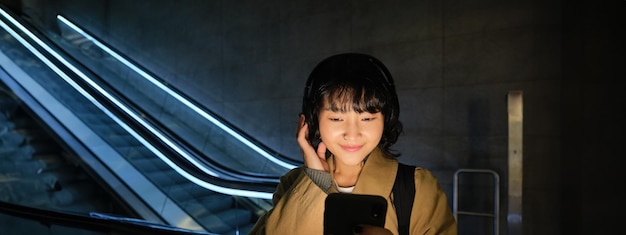 무료 사진 헤드폰을 입은 아름다운 한국 소녀의 초상화 에스컬레이터 근처에 서서 집으로 돌아갑니다