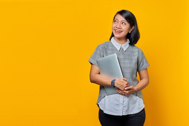 Портрет красивой улыбающейся женщины, держащей ноутбук и смотрящей в сторону, изолированной на желтом фоне
