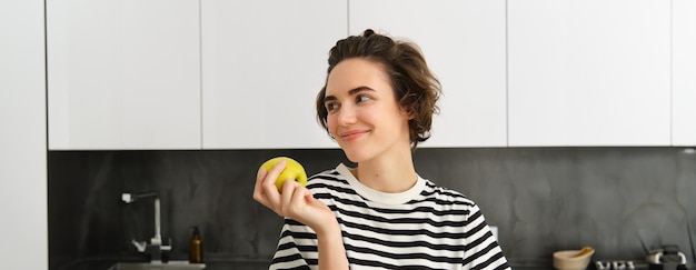 Бесплатное фото Портрет красивой улыбающейся молодой женщины, держащей яблоко, ест фрукты на кухне и выглядит счастливой