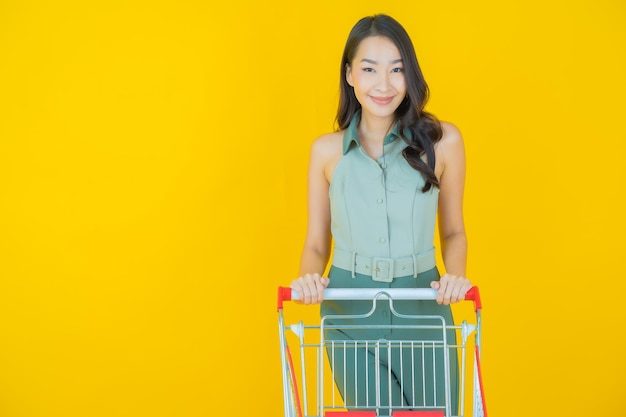 Портрет красивой молодой азиатской женщины улыбается с продуктовой корзиной из супермаркета на желтой стене