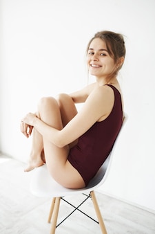 Портрет красивой молодой нежной женщины в усаживании swimwear усмехаясь назад на стуле поворачивая над белой стеной.