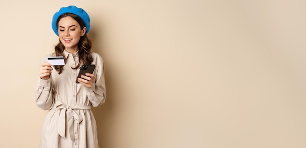 Бесплатное фото Портрет красивой молодой женщины 25 лет, использующей мобильный телефон для покупок с помощью кредитной карты, заказ доставки, покупка, стоящая на бежевом фоне