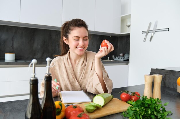 Бесплатное фото Портрет красивой молодой женщины на кухне, записывающей рецепт приготовления, сидя рядом с нарезкой