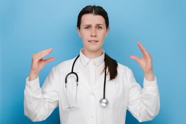 Портрет скучающей женщины-доктора в медицинском белом халате, показывающей жест бла-бла-бла руками и закатывающими глазами, позирует изолированной на синей стене фона студии. пустые обещания, лжец и концепция бла