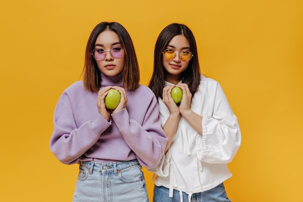 カラフル​な​サングラス​と​スウェットシャツ​の​ブルネット​の​若い​アジア​の​女の子​の​肖像画​は​、​孤立した​オレンジ色​の​壁​に​緑​の​新鮮な​リンゴ​を​保持します​。