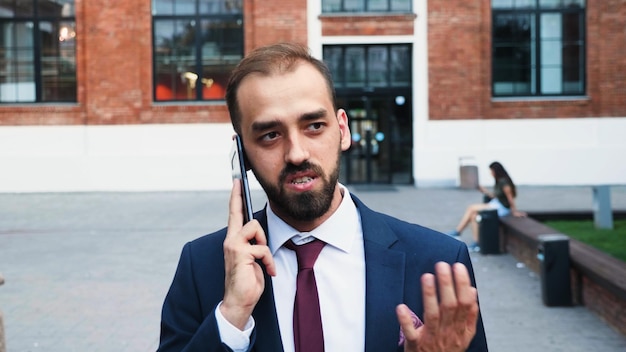Портрет бизнесмена в дипломатическом костюме, стоящего снаружи впереди на запускающем офисе, разговаривает на смартфоне, обсуждая финансовую стратегию с удаленным коллегой. концепция на открытом воздухе