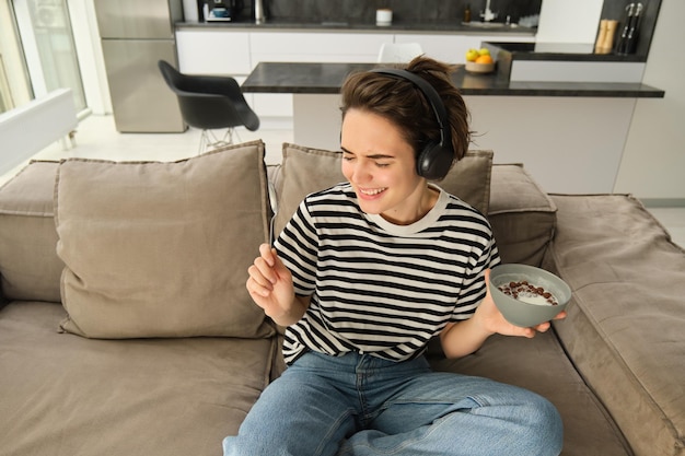 Бесплатное фото Портрет беззаботной улыбающейся женщины слушает музыку в наушниках, держащей миску с хлопьями