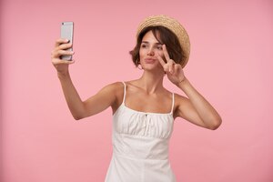 Бесплатное фото Портрет очаровательной молодой женщины с короткими каштановыми волосами в белом летнем платье и соломенной шляпе, поднимающей руку с жестом мира, делая селфи со своим смартфоном, изолированные