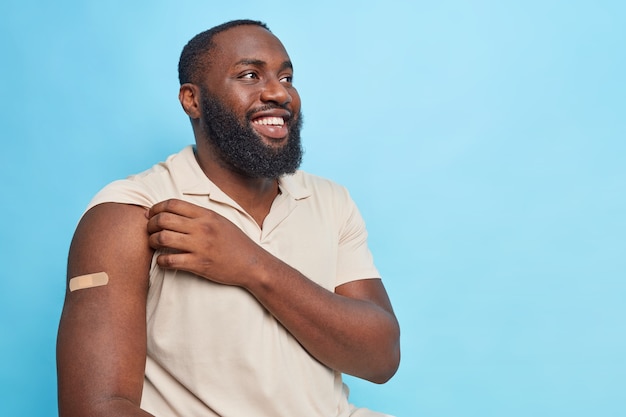 Портрет жизнерадостного бородатого афроамериканца показывает руку с гипсовой повязкой, счастливой получить вакцинацию, смотрит в сторону и улыбается, одетый в повседневную футболку, изолированную над синей стеной