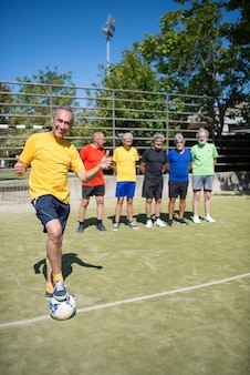 サッカー場​の​陽気な​年配​の​男性​の​肖像画​。​立っている​、​ボール​を​蹴る​、​バックグラウンド​で​チームメイト​の​スポーツ服​を​着た​白髪​の​チームリーダー​。​サッカー​、​スポーツ​、​レジャー​の​コンセプト