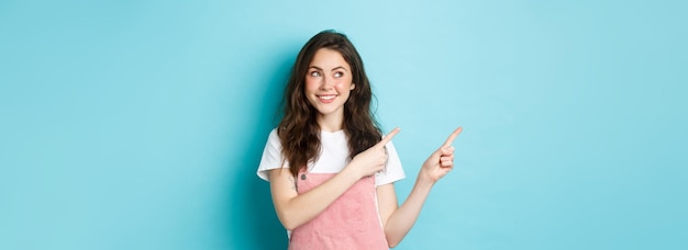 Бесплатное фото Портрет веселой милой девушки в розовом комбинезоне и белой футболке, смотрящей на продукт, указывающей пальцем