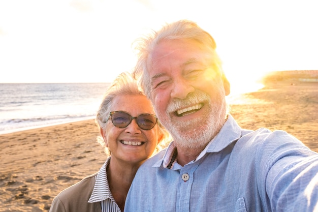 背景の夕日と一緒に自分撮りを撮ってカメラを見ているビーチで夏を楽しんでいる成熟した老人のカップルの肖像画。屋外を旅行する2人のアクティブな先輩。 Premium写真