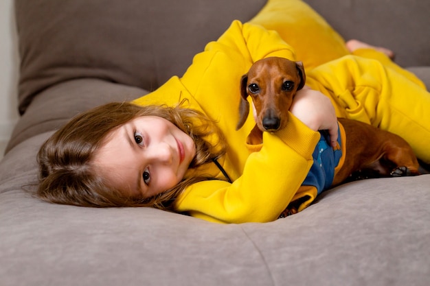 Портрет милой маленькой девочки в желтой одежде, лежащей в постели с карликовой таксой в синем комбинезоне
