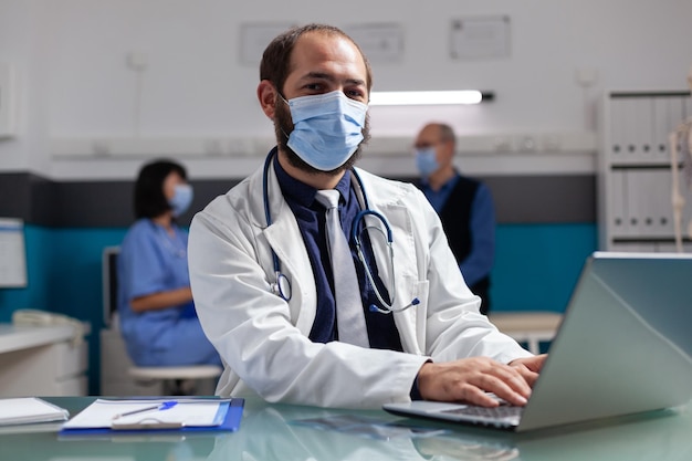 Бесплатное фото Портрет доктора, смотрящего в камеру и использующего компьютер в офисе. врач в маске для лица в белом халате на консультации, работающий с ноутбуком для медицинской карьеры.