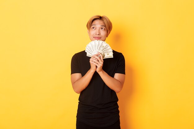 Портрет мечтательного красивого азиатского парня, показывающего свои сбережения и размышления, смотрящего в левый верхний угол, держащего деньги, стоящего у желтой стены