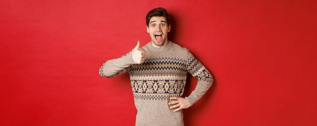 クリスマスセーターの興奮した、魅力的な男の肖像画、親指を立てて驚いて見える、良いことをお勧めし、クリスマス関連製品を宣伝します。