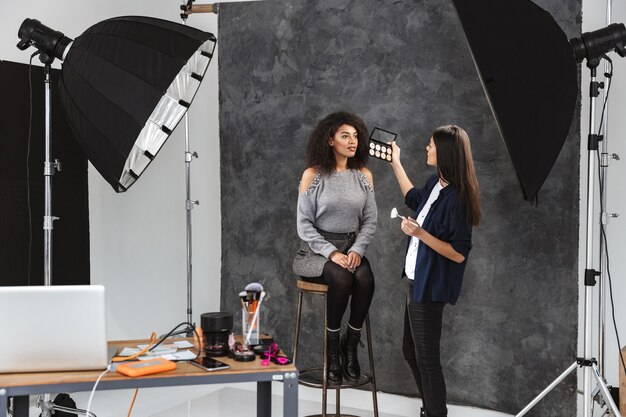 Портрет визажиста, наносящего косметику женщине во время фотосессии с профессиональной камерой и софтбоксом в студии