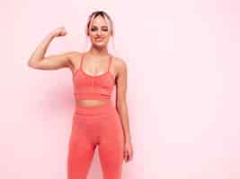 Бесплатное фото Портрет фитнес-женщины в розовой спортивной одежде. молодая красивая модель с идеальным телом. женщина позирует у стены в студии. веселая и счастливая.