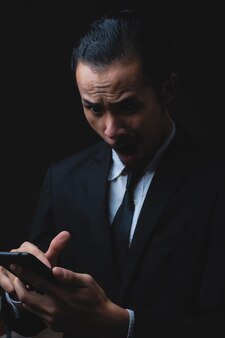 Портрет разочарованного кавказского молодого бизнесмена и предпринимателя, гримасничающего, глядя на телефон в строгой одежде на темном черном фоне