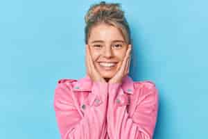 무료 사진 잘 생긴 여성의 미소의 초상화는 긍정적으로 뺨에 손을 대고 있고 건강한 탈지색 치아가 파란색 배경 위에 격리된 분홍색 재킷을 입고 즐거운 것을 듣습니다. 행복한 감정 개념