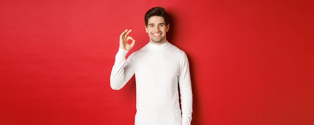 Бесплатное фото Портрет счастливого и довольного красивого мужчины в белом свитере, показывающего хороший знак и улыбающегося, одобряющего что-то хорошее, рекомендующего магазин, стоящего на красном фоне.