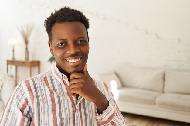 Бесплатное фото Портрет счастливого позитивного молодого афро-американского мужчины, сидящего в помещении с рукой на подбородке