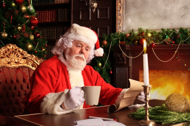 크리스마스 트리 근처 집에 있는 자신의 방에 앉아 크리스마스 편지나 위시리스트를 읽는 행복한 산타클로스의 초상화.