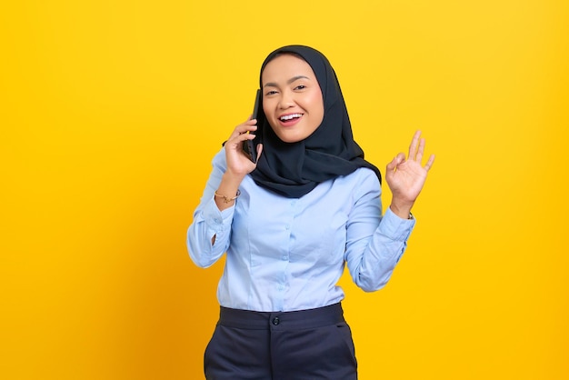 Портрет счастливой молодой азиатской женщины разговаривает по мобильному телефону и жестикулирует хорошо знаком на желтом фоне