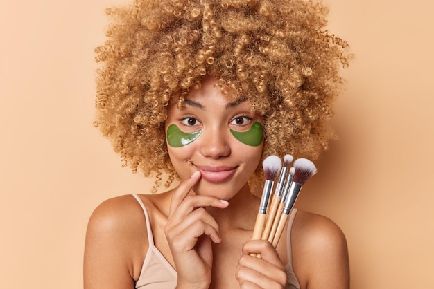 無料写真 喜んでいる縮れ毛の女性の肖像画は、目の下に緑色のハイドロゲルパッチを適用してしわを取り除き、茶色の背景の上に孤立した化粧をするための化粧ブラシを保持します女性の美容美容