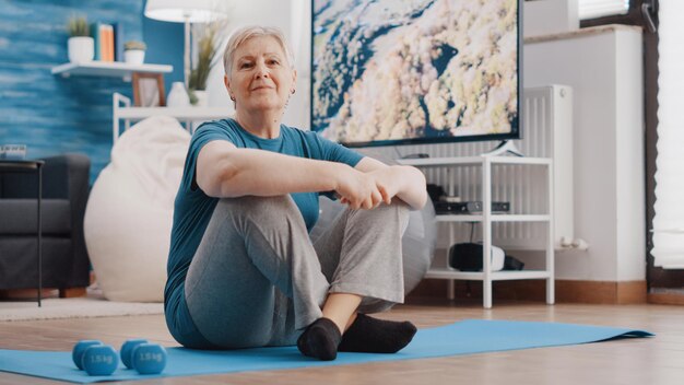 집에서 신체 운동과 운동 훈련을 준비하는 고위 여성의 초상화. 은퇴한 사람은 요가 매트에 앉아 카메라를 보고 피트니스 장비를 사용할 준비가 되었습니다.