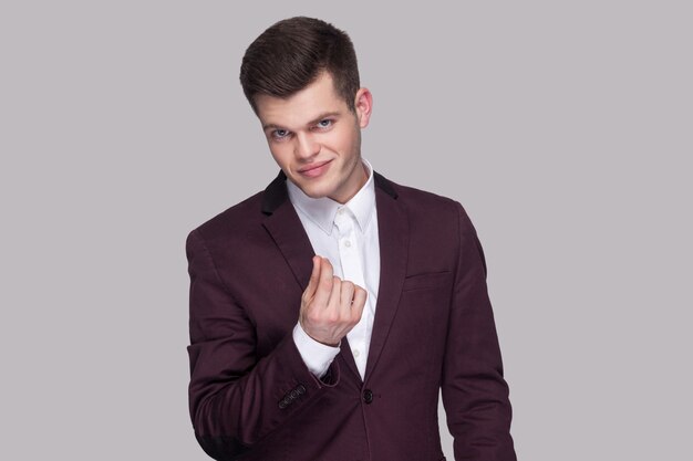 Портрет серьезного красивого молодого человека в фиолетовом костюме и белой рубашке, стоящего, смотрящего в камеру и показывающего денежный жест. крытая студия выстрел, изолированные на сером фоне.
