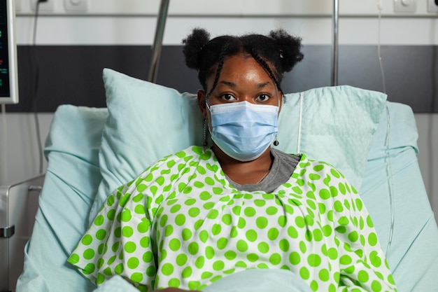 病棟での手術後に回復するコロナウイルス感染を防ぐために保護フェイスマスクを着用して、診察中にベッドに横たわっている病気の若い患者の肖像画。医療サービス