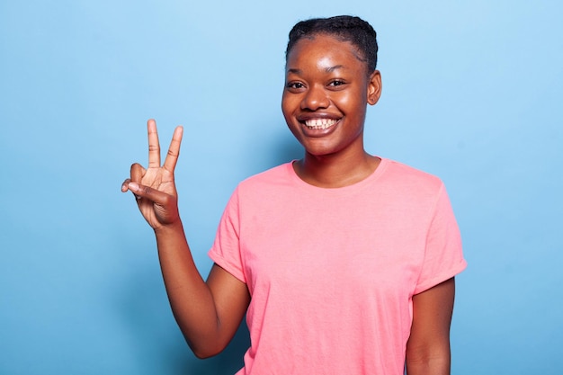 青い背景のスタジオに立っているカメラで指で平和のシンボルを作る笑顔のアフリカ系アメリカ人の若い女性の肖像画。楽しんで笑っているうれしそうなティーンエイジャー。お祝いのコンセプト