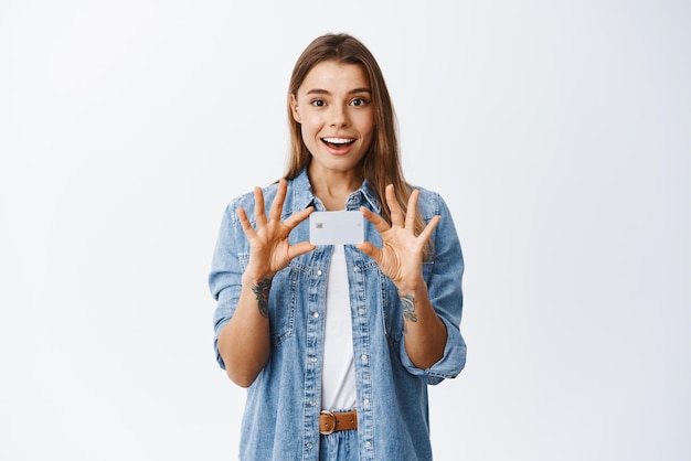 Бесплатное фото Портрет улыбающейся и удивленной молодой женщины, держащей пластиковую кредитную карту в руках над сундуком, рекламирующей новую банковскую функцию, рекомендующую банк на белом фоне