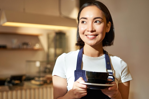 無料写真 カフェで働くドリンクのカップを保持しているコーヒー ウェイトレスを提供する制服を着た笑顔のアジアのバリスタの女の子の肖像画