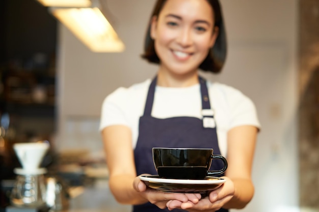 無料写真 一杯のコーヒーを提供するカフェの制服を着た笑顔のアジアの女の子のバリスタ ウェイトレスの肖像画は、dri を準備します