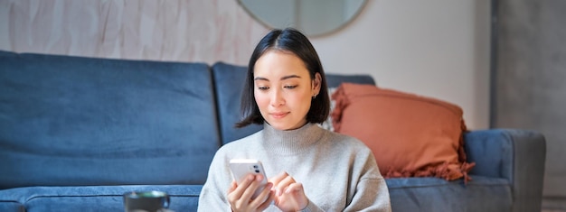 무료 사진 집에 앉아 웃고 있는 아시아 여성의 초상화는 스마트폰 앱을 이용해 누군가에게 배달 메시지를 주문한다