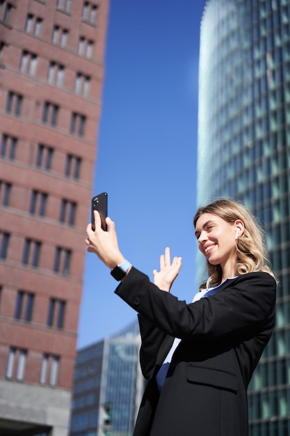 Бесплатное фото Портрет улыбающейся корпоративной женщины по видеозвонку на улице, держащей мобильный телефон и машущей смартфо