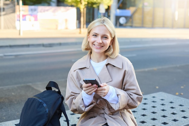 無料写真 友人の注文を待っているスマートフォンで通りベンチに座っている笑顔の女性モデル ⁇