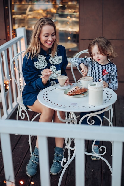 無料写真 カフェで彼女の娘とコーヒーを楽しんでいるアヒルと青いブーツと青いドレスで色白の髪の幸せな女性の笑顔の肖像画。屋外のテーブルで母親のそばに座ってカカオをかき混ぜる素敵な女の子。