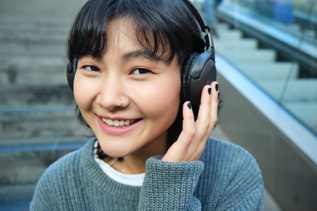 無料写真 ヘッドフォンで笑顔の韓国の女の子の肖像画は、スマートフォンを使用し、モールの階段に座ってビデオを見る