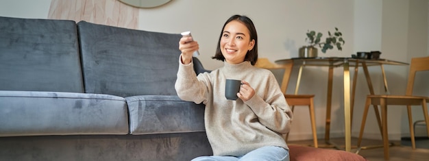 무료 사진 리모컨을 들고 채널을 전환하는 동안 tv 근처에 앉아 웃고 있는 한국 여성의 초상화